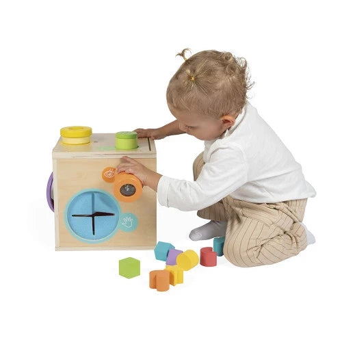 Toddler interacts with a wooden sensory cube and colorful blocks featuring yellow, green, blue, and orange elements.