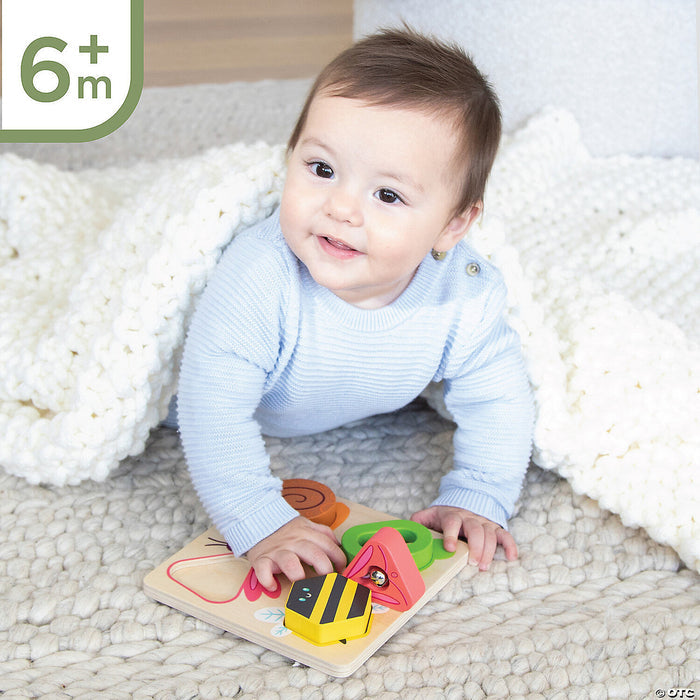 An infant plays with a wooden sensory puzzle featuring colorful shape and bug pieces on a white knit rug.
