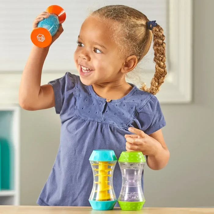 A child plays with three hourglass-shaped tubes featuring blue glitter, a yellow spiral, and floating purple beads.