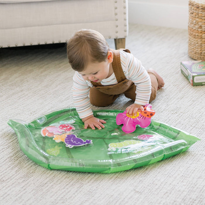 Baby pressing hands on a green leaf-shaped inflatable mat with water and floating colorful animal shapes inside.