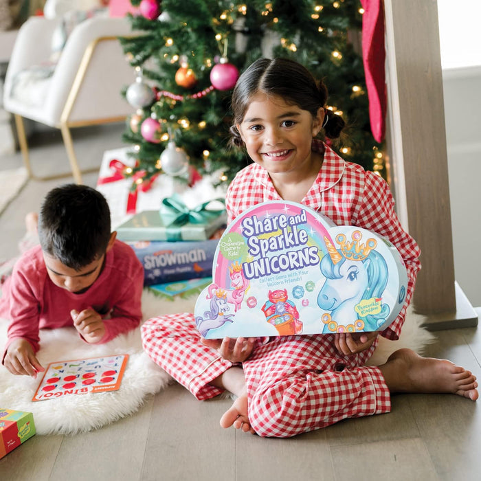 A girl in red pajamas holds a cloud-shaped game box featuring colorful unicorns and gems near a Christmas tree.