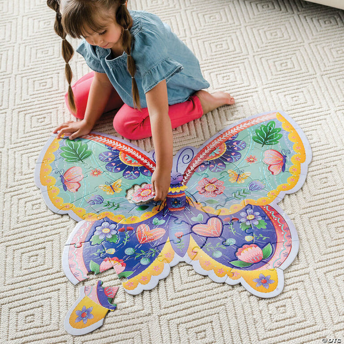 A girl sits on a rug assembling a large butterfly-shaped floor puzzle featuring vibrant floral and heart designs.