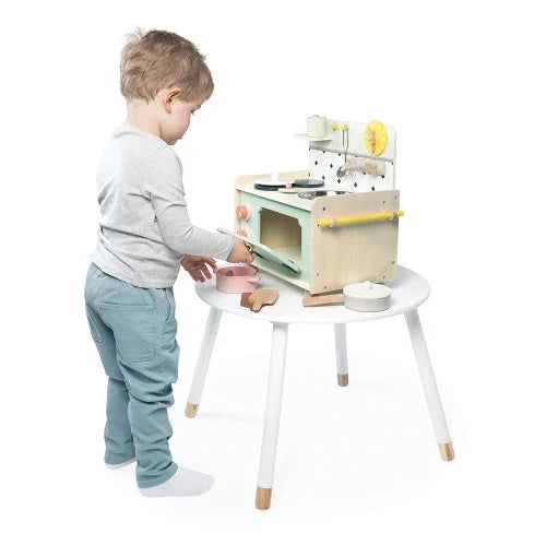 Young boy plays with a mint and wood toy kitchenette on a round white table with toy pots and pans.
