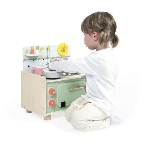Toddler playing with a pastel green and wood toy kitchen featuring an oven, stovetop, and mini cookware.
