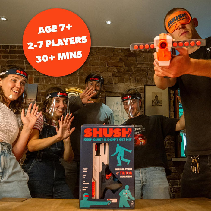 Friends wear face shields playing a party game. A blindfolded man aims a foam blaster over the blue and orange game box.