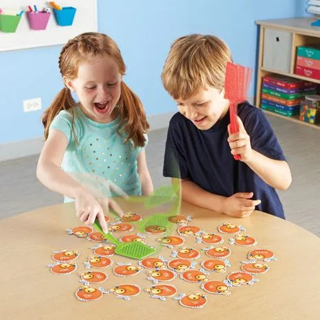 Two children use green and red fly swatters to swat orange fly-shaped word cards on a wooden table.