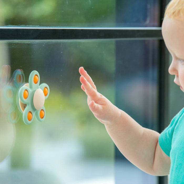 Mint green flower-shaped toy with five orange centers suctioned to glass as a toddler reaches out to touch it.