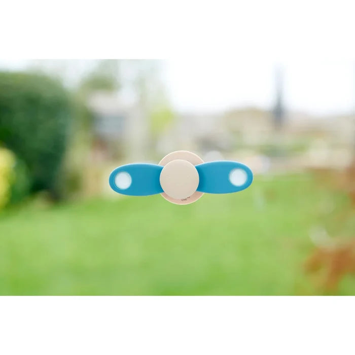 Light blue and cream two-blade propeller spinner suctioned to a window with a blurred green garden background.
