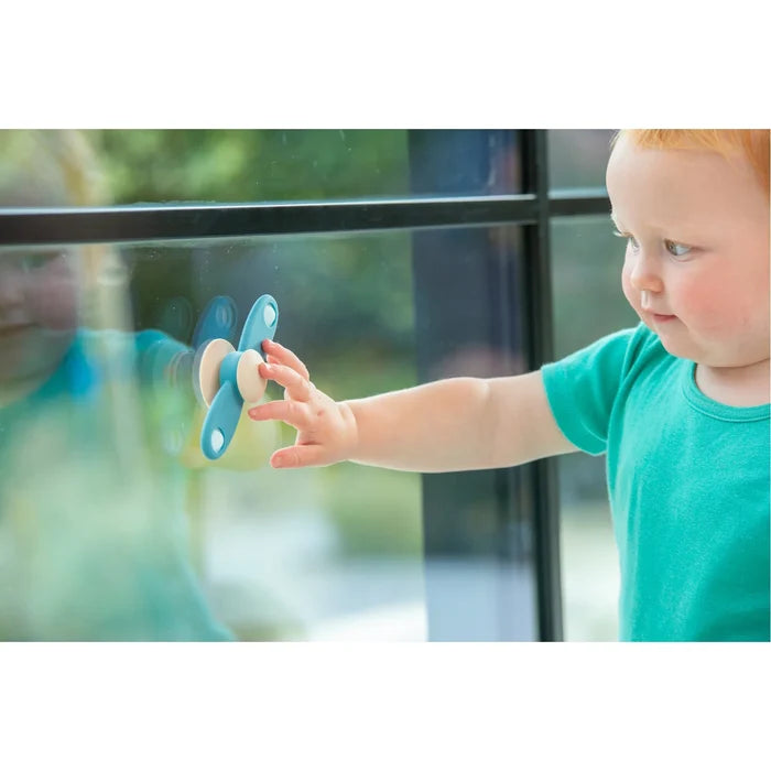 Toddler spins a blue and beige silicone propeller toy suctioned to a glass window.