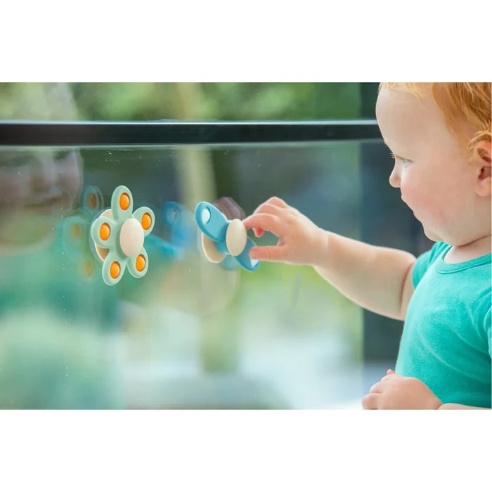 A toddler playing with mint and blue suction cup spinners in flower and propeller shapes on a glass window.