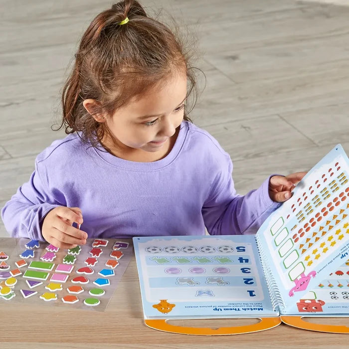 A girl playing with a spiral activity book and colorful sticker sheets featuring shapes, numbers, and animals.