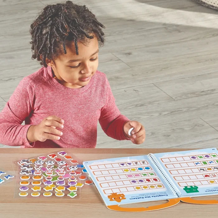 A young child uses colorful stickers to complete pattern activities in a spiral-bound book on a wooden table.