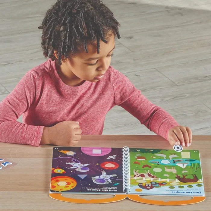 Child in red shirt placing a sticker in a spiral-bound shape activity book with space and farm-themed scenes.