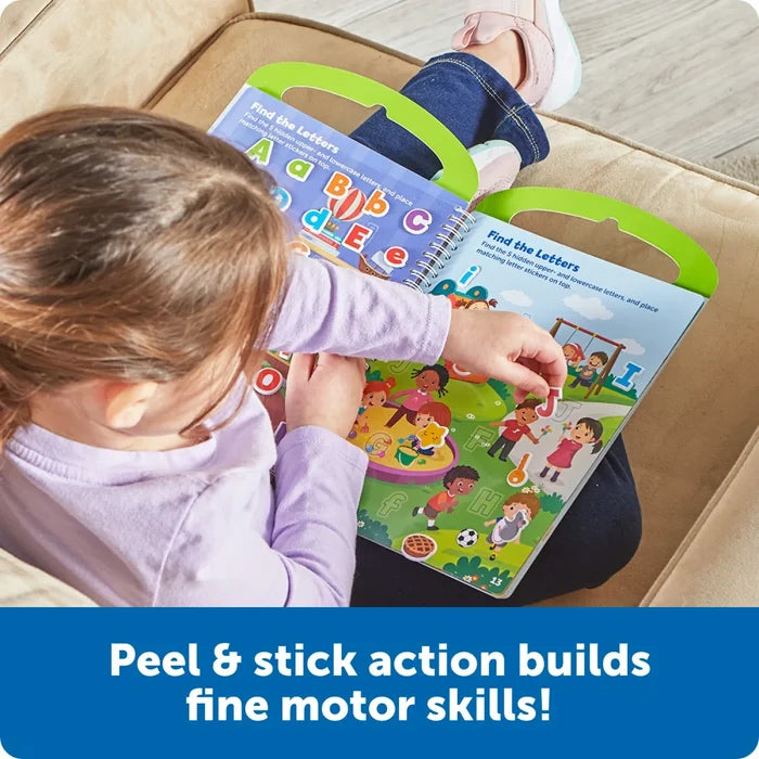 A child sticks a red letter sticker into a colorful alphabet activity book featuring playground scenes.