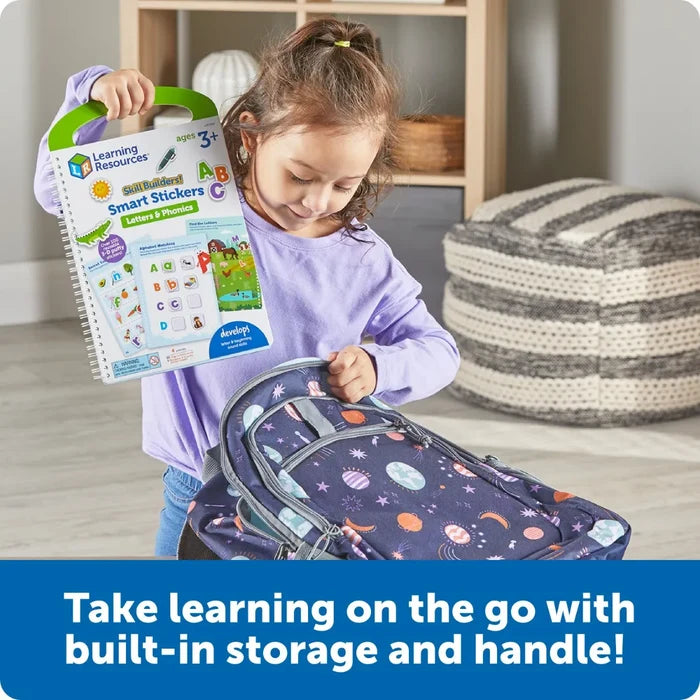 A child packs a white spiral-bound activity book with a green handle into a blue galaxy-print backpack.