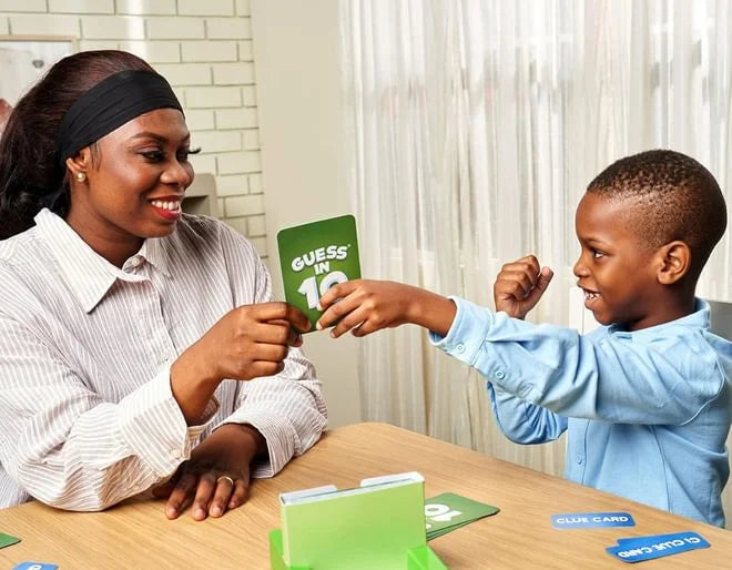 A woman hands a green Guess in 10 card to a cheering boy at a wooden table with a green box and blue clue cards.
