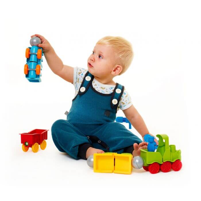 Toddler playing with colorful, chunky magnetic train pieces including a green engine, red wagon, and blue car.