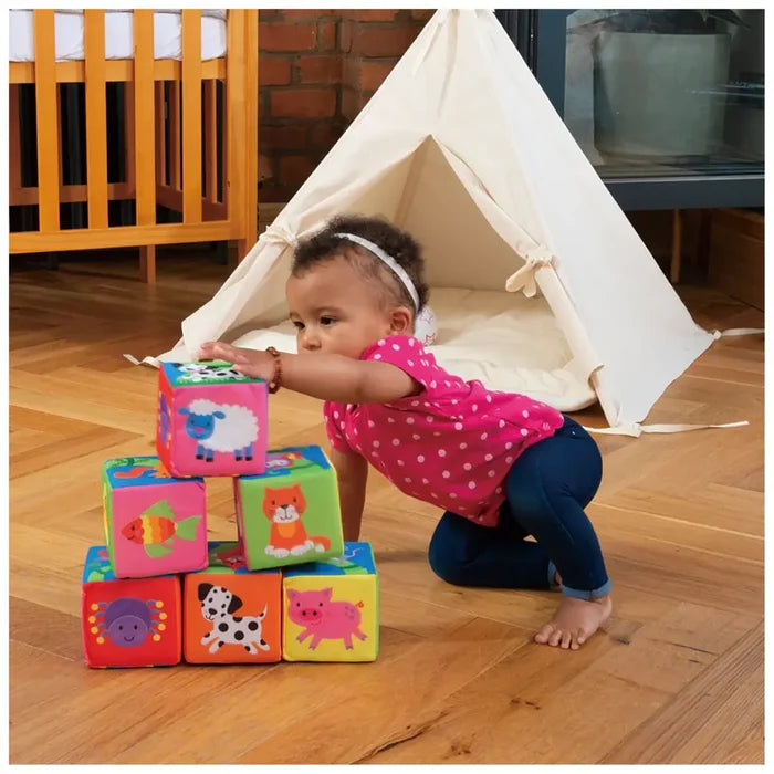 Six colorful soft blocks with animal drawings stacked in a pyramid as a baby reaches for the top cube.