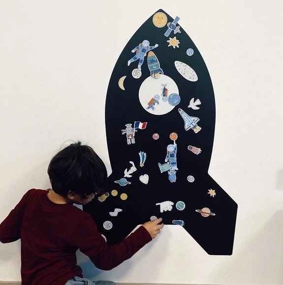 Child places space-themed magnets onto a large black rocket-shaped silhouette mounted on a white wall.
