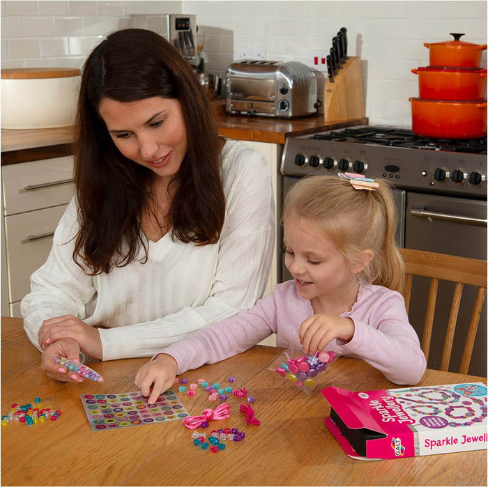Woman and child crafting with colorful beads and stickers from a pink Sparkle Jewellery box on a wooden kitchen table.
