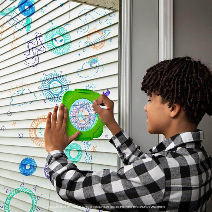 Child uses a green Spirograph tool to draw colorful geometric patterns on a window with markers.