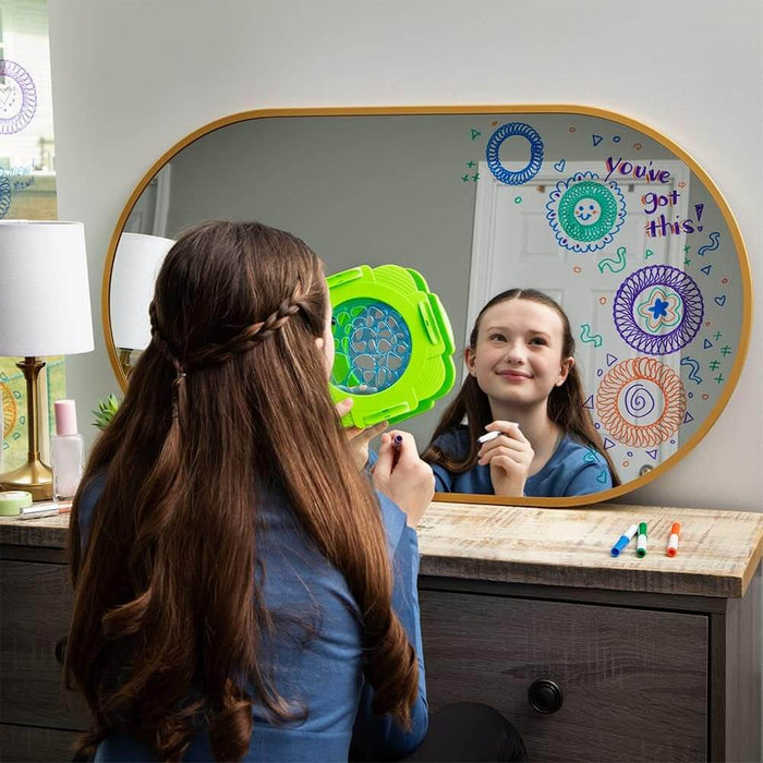 A girl uses a green Spirograph tool to draw colorful patterns and messages on a large oval vanity mirror.