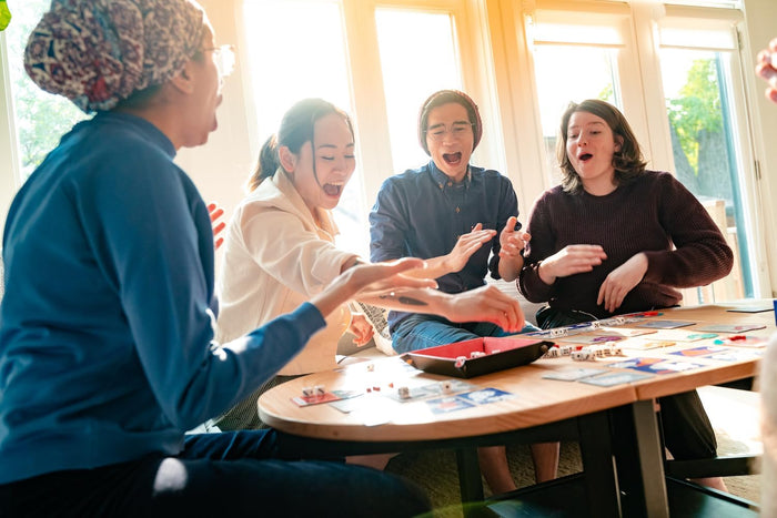 Four friends excitedly play a board game with white dice, cards, and a red tray on a wooden coffee table.
