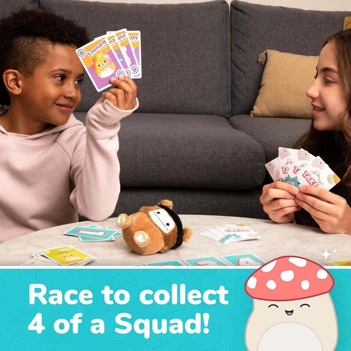 Two children play a Squishmallows card game with character cards and a small plush mascot on a table.