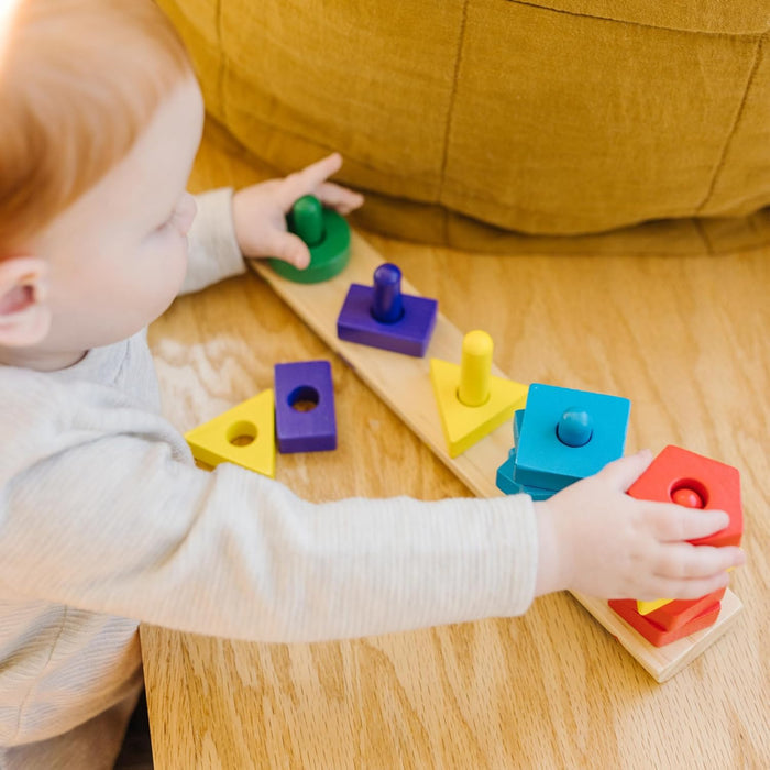 Toddler stacks colorful wooden circles, squares, and triangles onto a natural wood sorting board with five pegs.