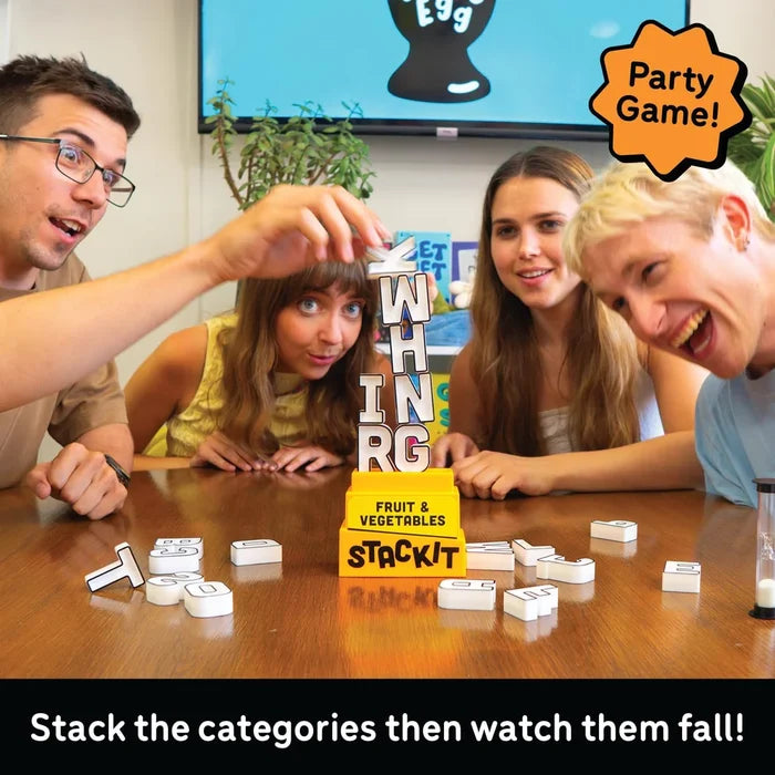 Four people laughing around a wooden table playing a fruit and vegetable category stacking game. White category tiles are scattered on the table, with one person carefully stacking the word 'FRUIT' vertically above a yellow 'FRUIT & VEGETABLES STACKIT' box.