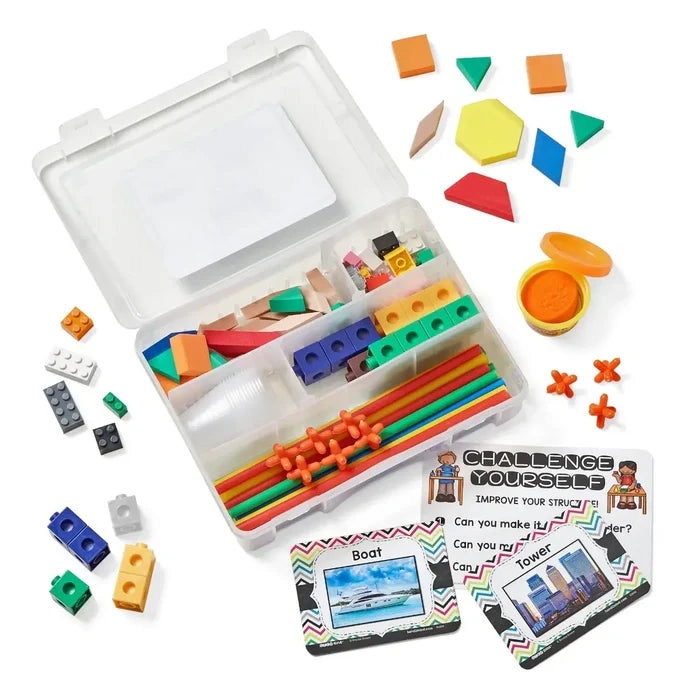 Clear plastic bin filled with colorful blocks, geometric shapes, straws, orange dough, and challenge cards.