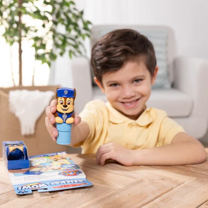 Smiling boy holds a blue dog-shaped sticker stamper over a PAW Patrol activity pad and box on a light wood table.