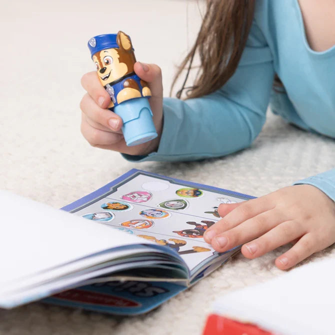 Child's hand holding a blue Chase character stamper over an activity pad with round PAW Patrol stickers.