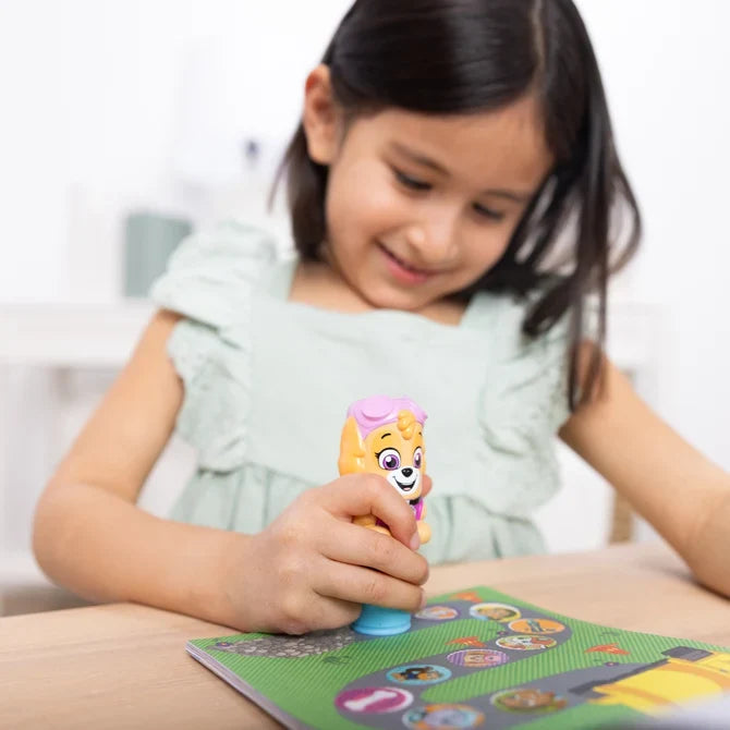 A girl smiles as she uses a pink Skye PAW Patrol sticker stamper to place stickers in a colorful activity book.