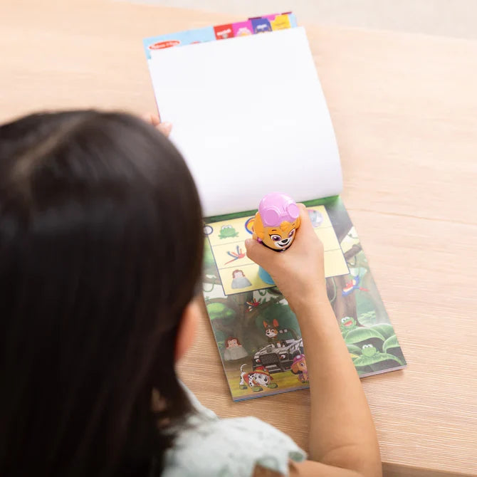 A child stamps a colorful PAW Patrol jungle page using a pink Skye-themed sticker stamper on a light wood table.