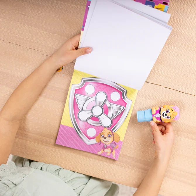 Child's hands using a blue Skye sticker stamper on a pink PAW Patrol activity pad with circular sticker targets.