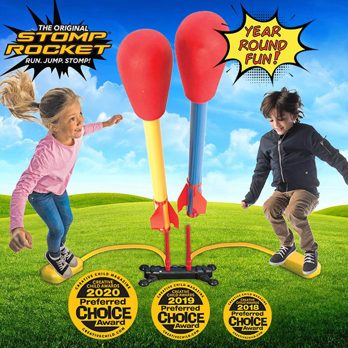 Two children jumping on yellow pads to launch red foam rockets with blue and yellow bodies into the sky.