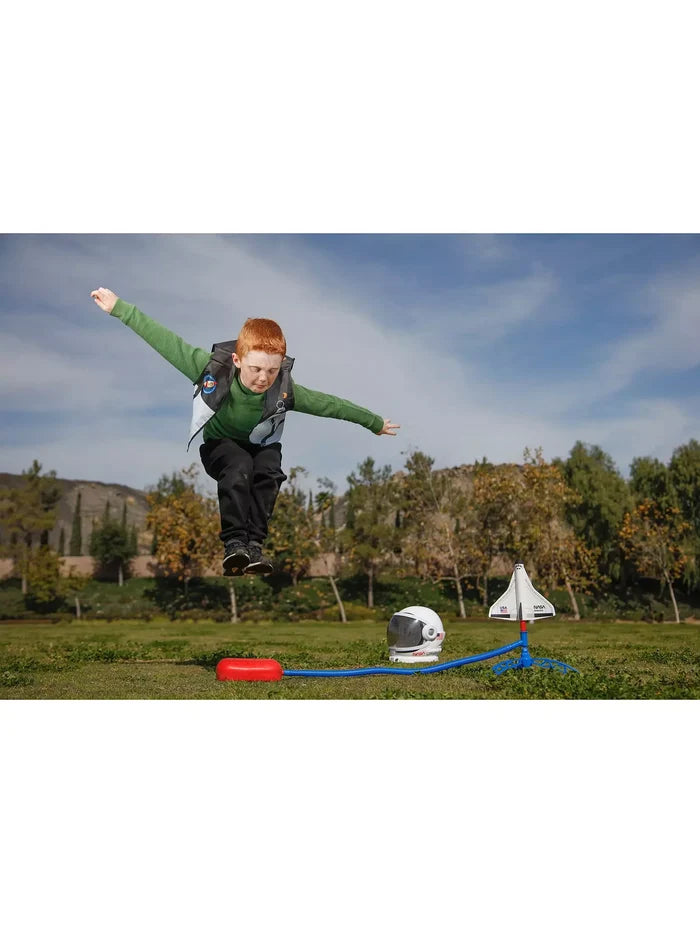 A child jumps on a red stomp pad to launch a white toy NASA shuttle near an astronaut helmet on a green lawn.