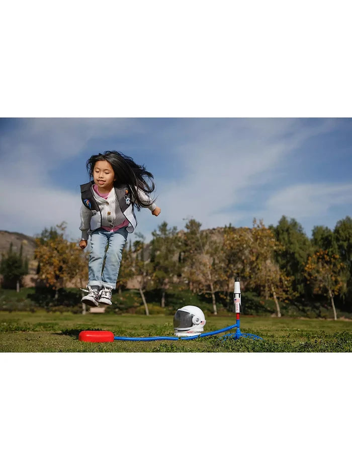 A girl jumps onto a red stomp pad to launch a NASA toy rocket in a park, with an astronaut helmet on the grass nearby.