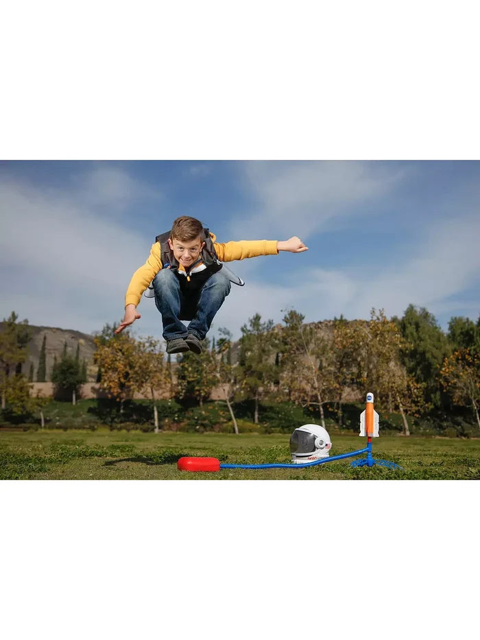 Boy jumps on a red stomp pad to launch an orange NASA rocket, with a toy astronaut helmet nearby on the grass.