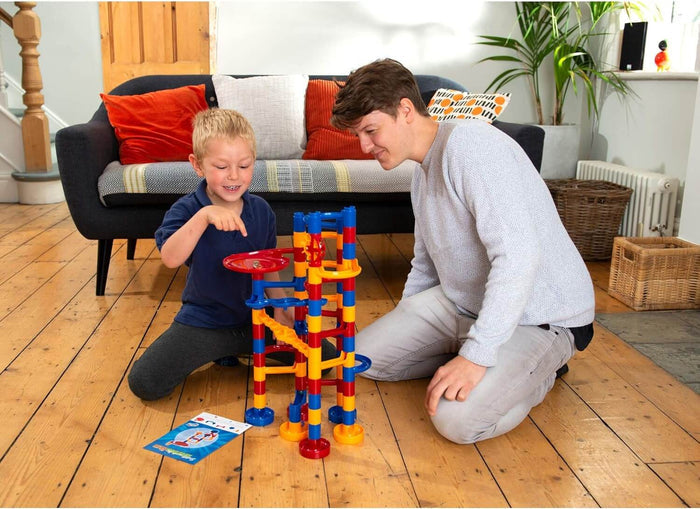 Man and child play with a tall red, blue, and yellow plastic marble run featuring spiraling tracks on a wooden floor.