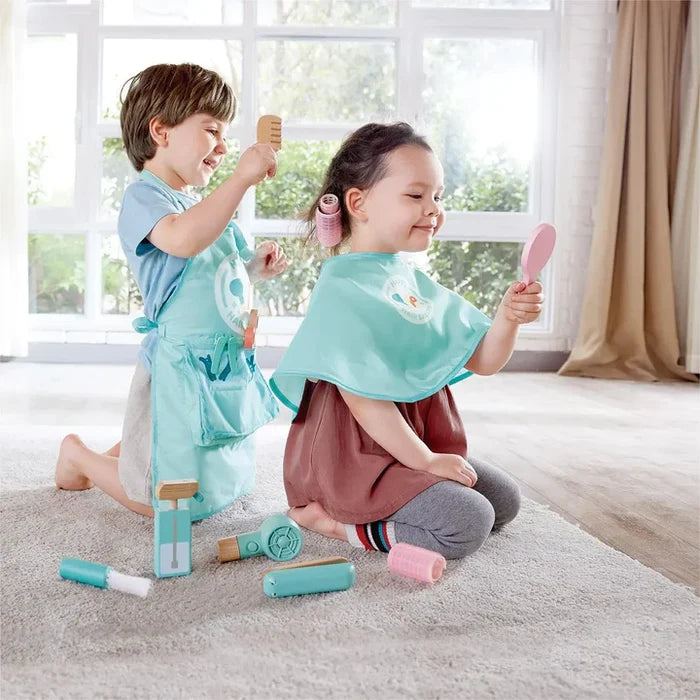 Kids playing with a wooden hair salon set including a blue apron, cape, hairdryer, comb, and pink mirror on a rug.