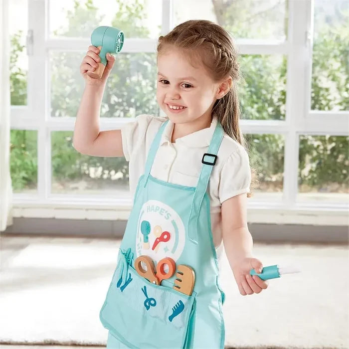 Child in a teal apron holding a toy hair dryer and wooden styling tools from a hair salon play set.