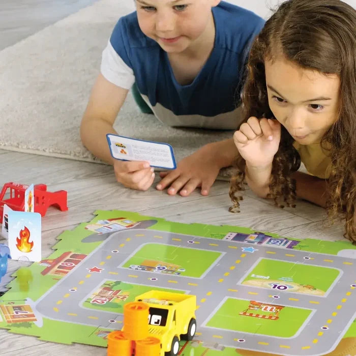 Two children play a city board game with a yellow toy truck, numbered buildings, and cards on a colorful map.