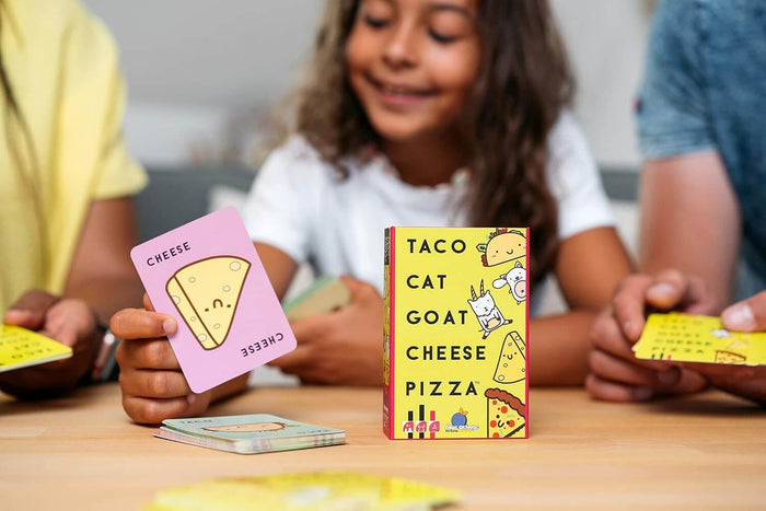 Hand holds a pink cartoon cheese card next to the yellow Taco Cat Goat Cheese Pizza game box on a wooden table.