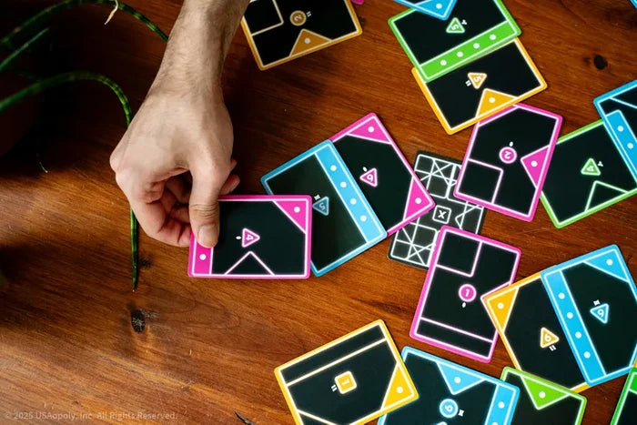 A hand places a neon-bordered black card on a wooden table among several other colorful geometric game cards.
