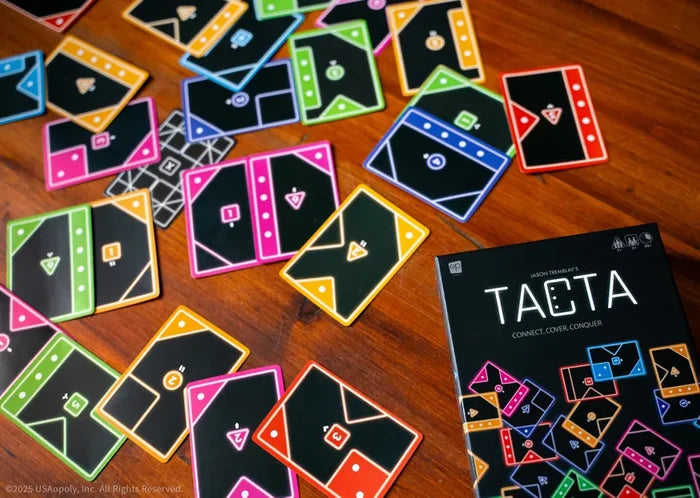 Black game box and cards with colorful neon geometric borders and icons scattered across a wooden table.