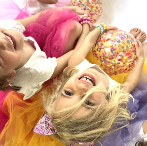 Clear round ball filled with bright multicolored confetti, held by children in colorful tutus.
