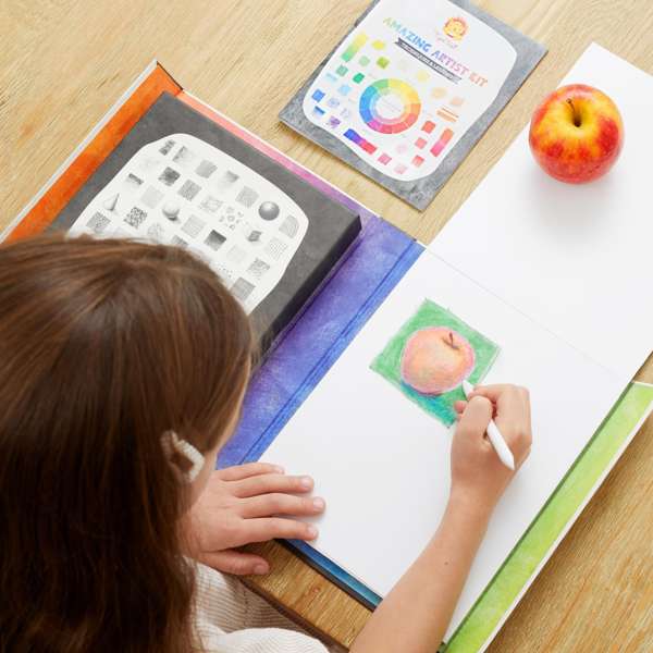 Child drawing a red apple using a white pencil from an art kit featuring shading guides and a color wheel booklet.