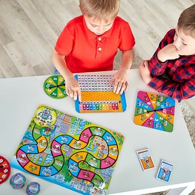 Two boys play an educational math game with a colorful winding board, spinners, and a multiplication grid.